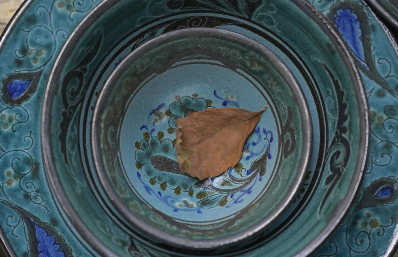 This close-up shows a traditional glazed ceramic bowl decorated primarily in turquoise, blue, black, and muted green tones. The scene documents Rishton ceramic craftsmanship. It documents cultural heritage in Fergana Reg. Фото: AIDEPCUL, CC BY 4.0, Викисклад