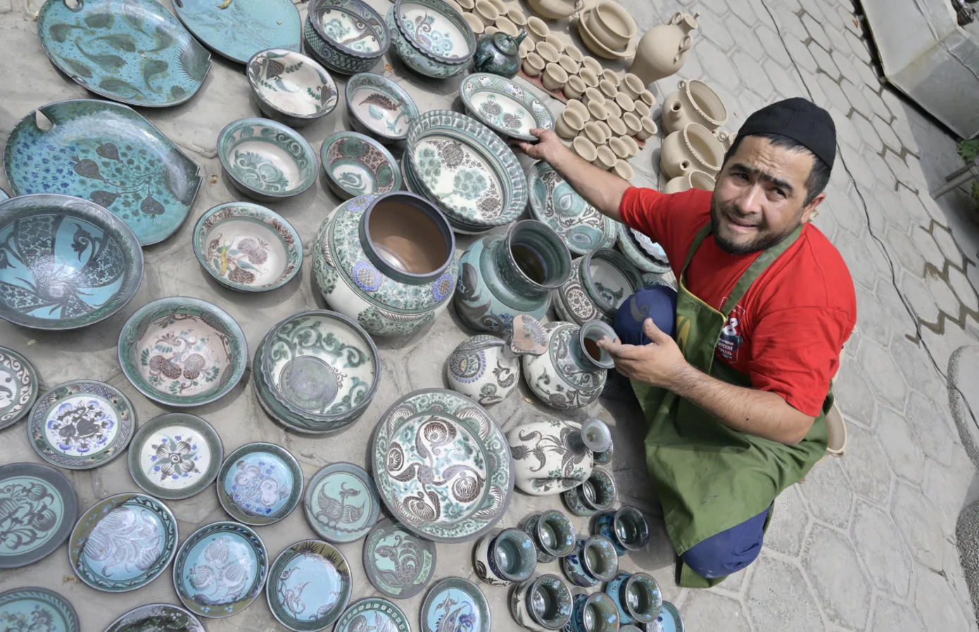 This overhead image presents a broad assortment of ceramic products arranged around a craftsman. The collection includes bowls, plates, jars, cups, teapots, and larger vessels. The scene documents Rishton ceramic craftsm. Фото: AIDEPCUL, CC BY 4.0, Викисклад