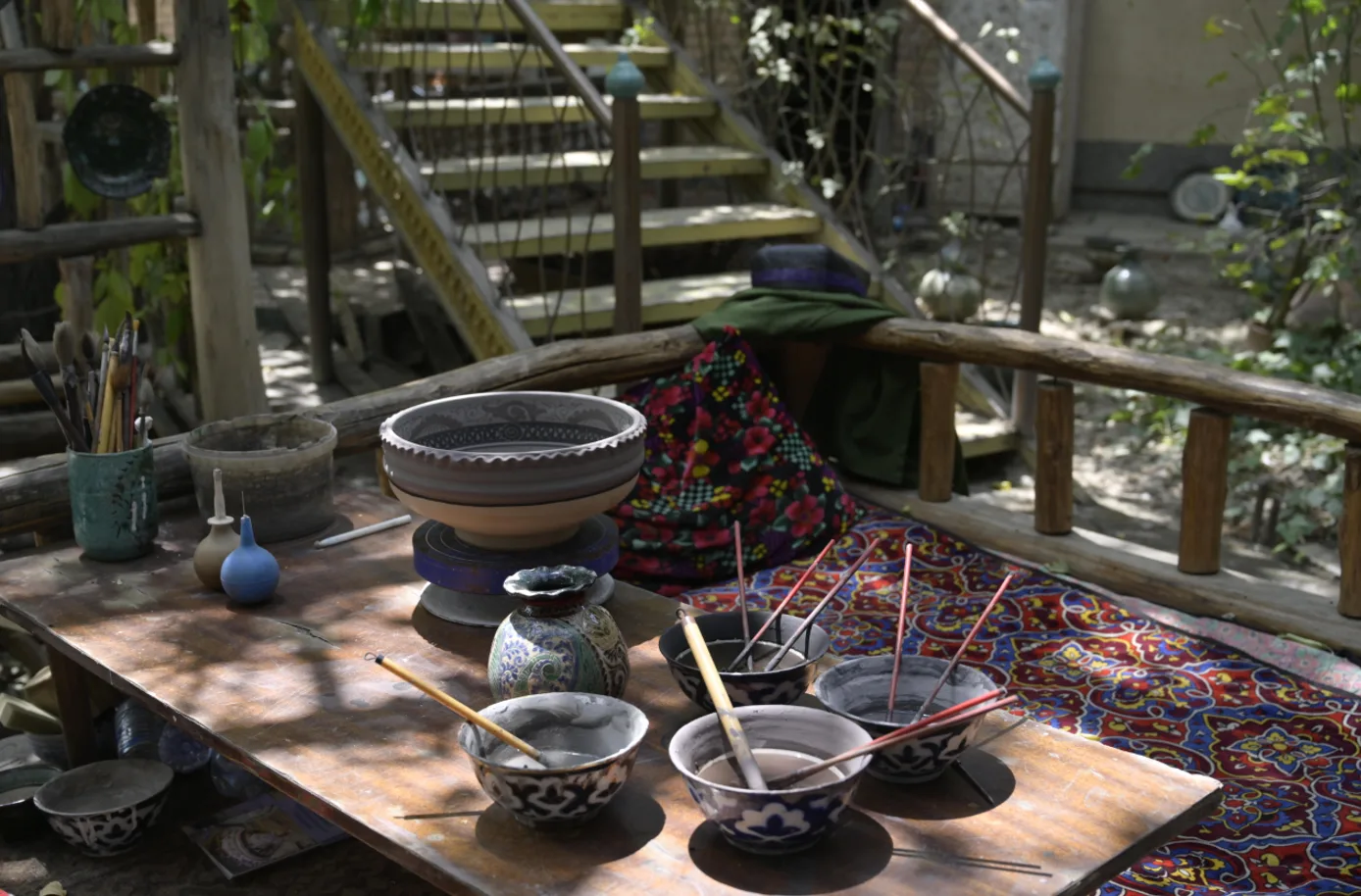 This image presents a traditional outdoor pottery workspace prepared for ceramic decoration. The scene documents Rishton ceramic craftsmanship. It documents cultural heritage in Fergana Region. The image records local tr. Фото: AIDEPCUL, CC BY 4.0, Викисклад